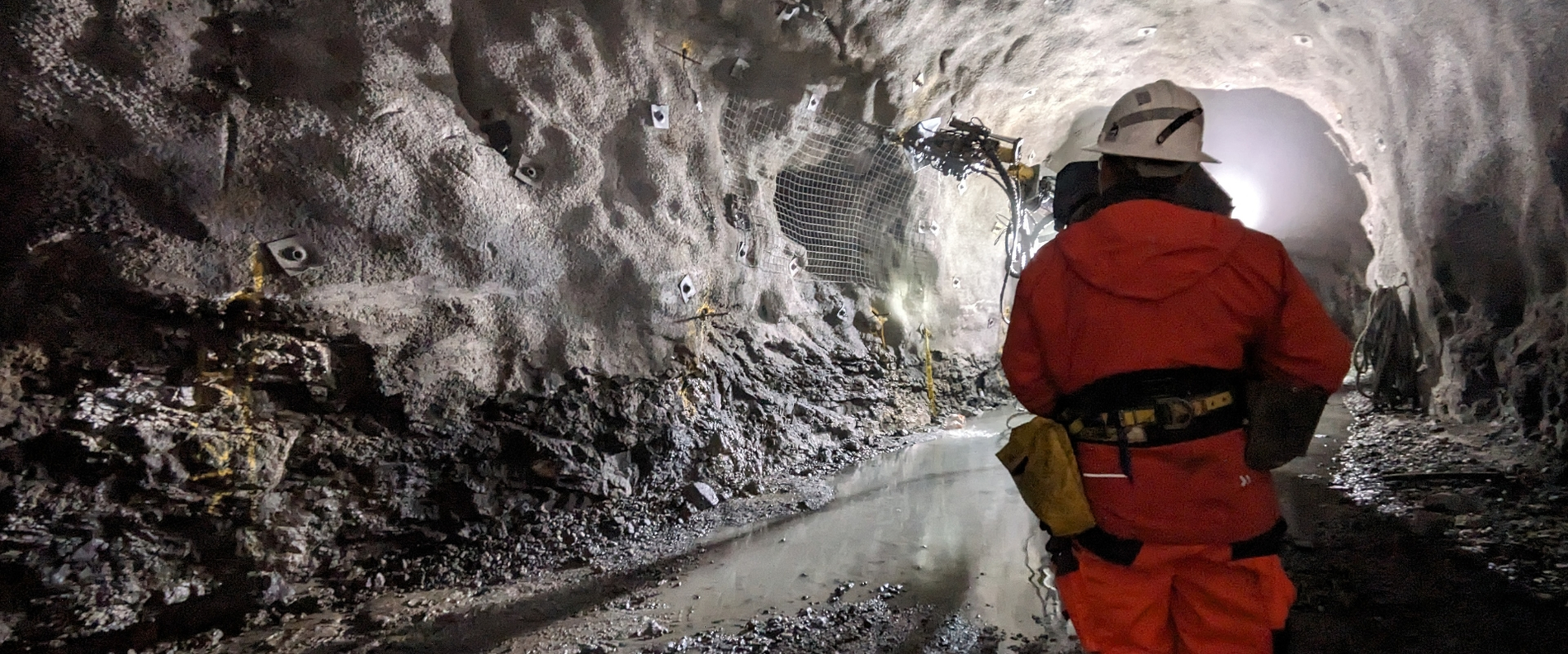Eine Person in orangefarbener Schutzkleidung steht in einem beleuchteten Tunnel, im Hintergrund sind Wasserpfützen, Steinanker und eine Bohrmaschine zu sehen.