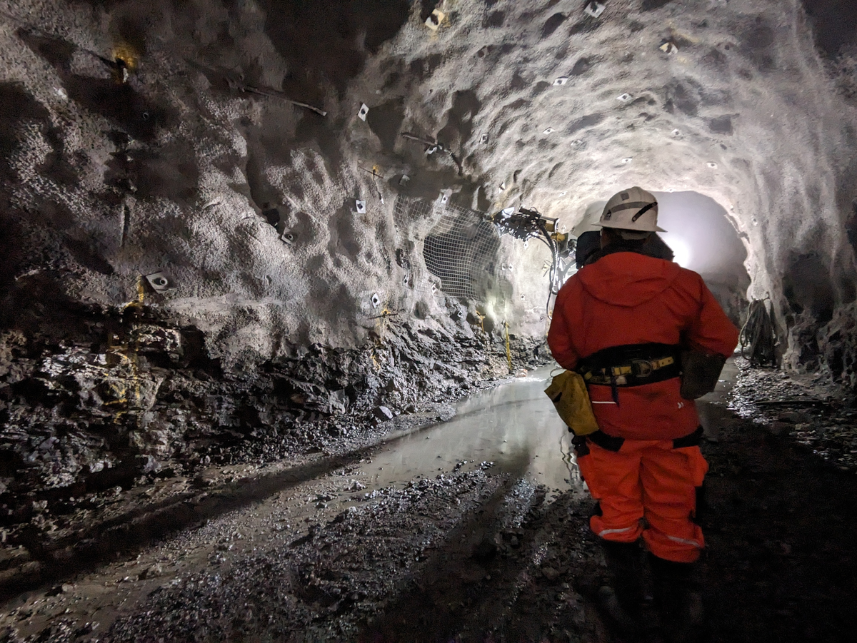 Eine Person in orangefarbener Schutzkleidung steht in einem beleuchteten Tunnel, im Hintergrund sind Wasserpfützen, Steinanker und eine Bohrmaschine zu sehen.